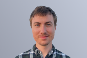 Portrait of a young man with short hair wearing a black and white plaid shirt, standing against a light gray background.