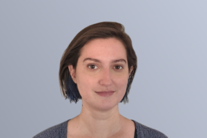 Portrait of a person with short, dark brown hair, wearing a gray cardigan and a hint of a smile against a light blue background.