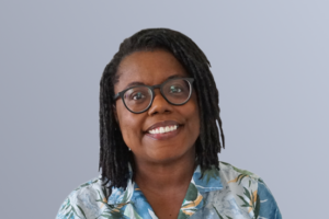 Portrait of a smiling person with glasses and dreadlocks, wearing a floral shirt against a plain background.