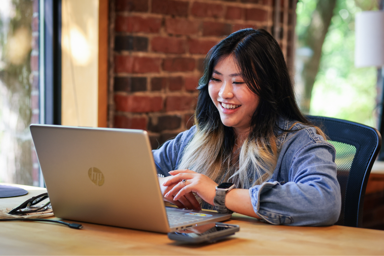 Person smiling and working on a laptop at a desk in a cozy, brick-walled room with natural light.