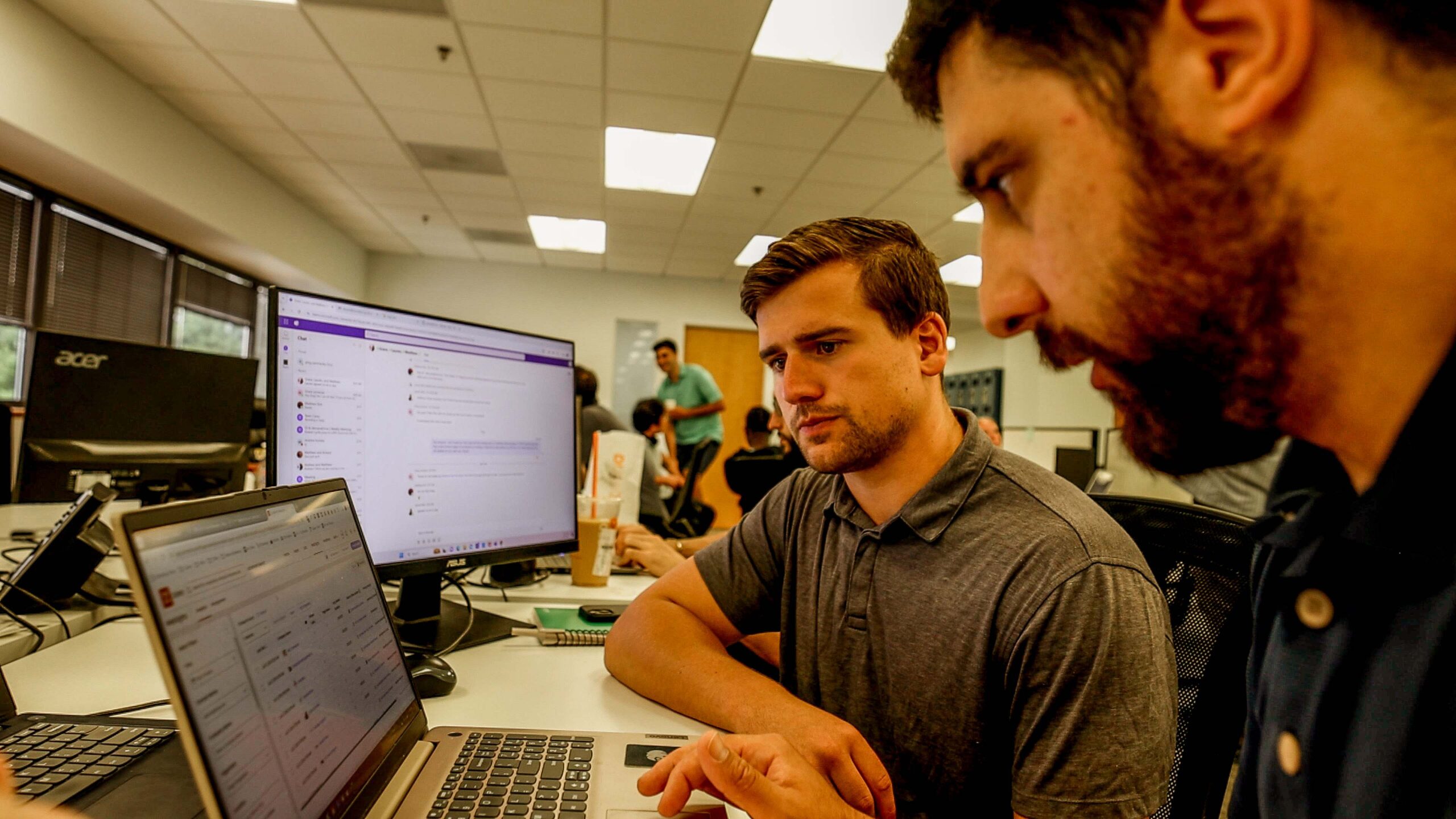 "Two men working on laptops at a desk in an office, screens displaying code and projects, with colleagues in the background"