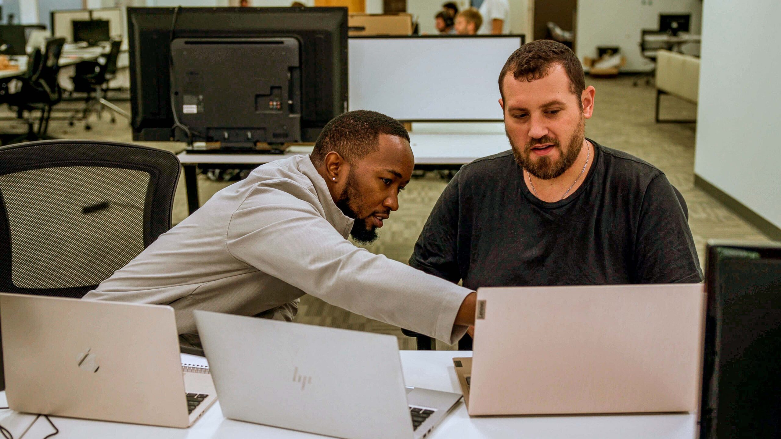 Two men collaborating over work at a desk in a modern office setting with laptops in front of them.